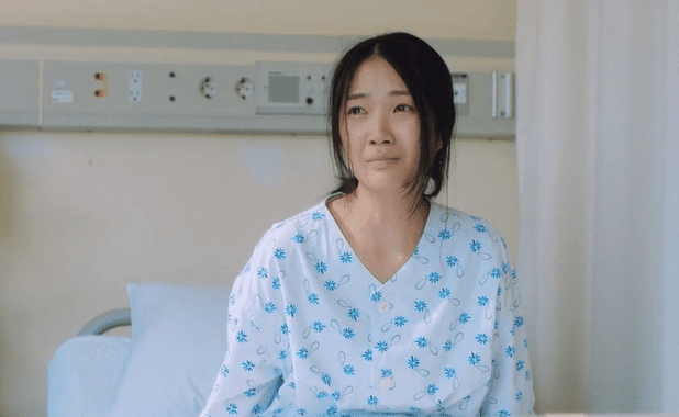 Mujer con bata de hospital sentada en una cama mostrando una expresión de preocupación en un entorno clínico