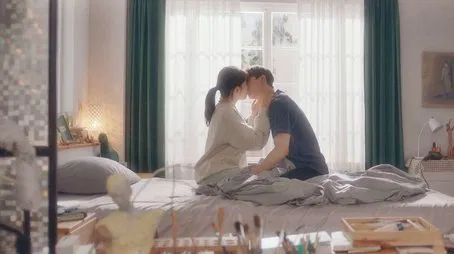 Pareja abrazándose en una cama cerca de una ventana, lo que sugiere un momento romántico.