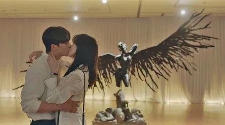 Pareja besándose apasionadamente en una galería de arte, con una gran escultura oscura alada en el fondo.
