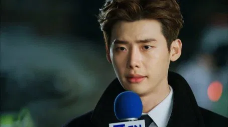 El actor coreano Lee Jong-suk con un traje oscuro, hablando por un micrófono.