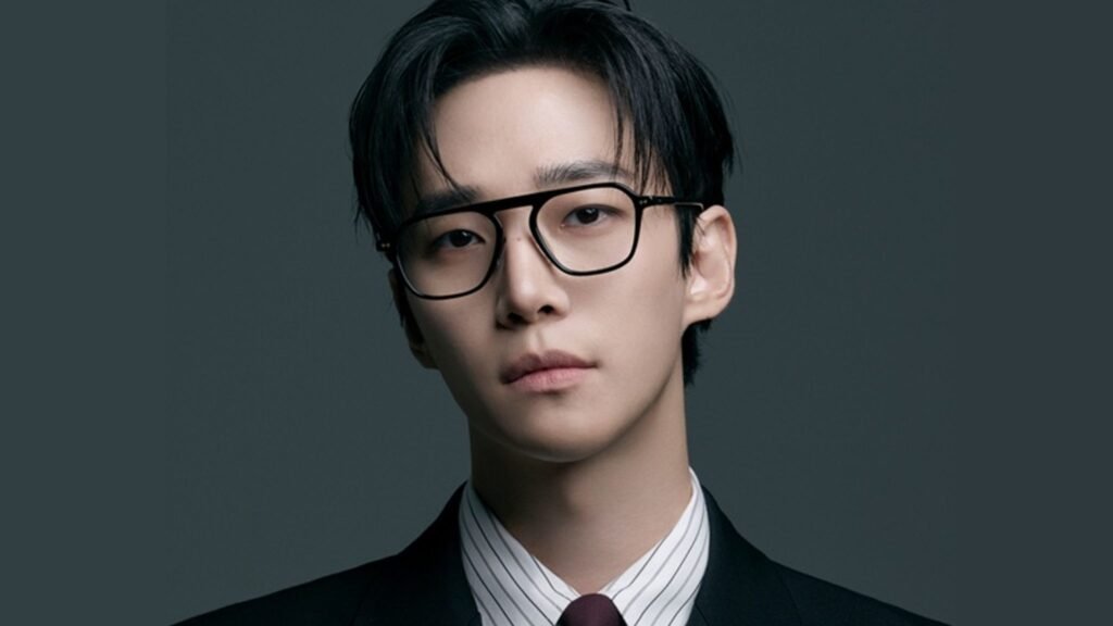 Primer plano del actor Lee Jun-ho con gafas de montura negra y traje formal mirando a cámara sobre fondo gris.