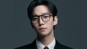 Primer plano del actor Lee Jun-ho con gafas de montura negra y traje formal mirando a cámara sobre fondo gris.